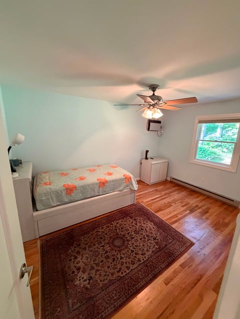 Small bedroom with white walls, wood floor, bed with orange patterned comforter, burgundy rug, ceiling fan with light, and window with green view outside.