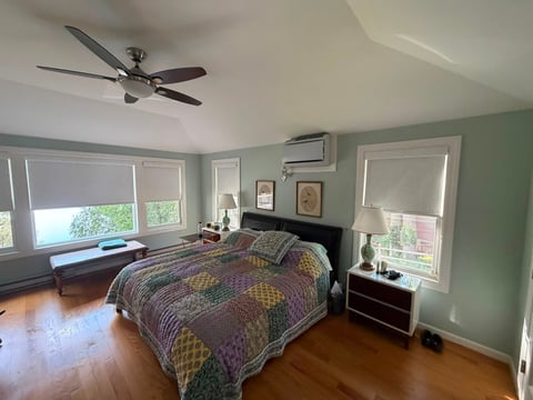 Bright bedroom with green walls, queen bed with colorful patterned bedspread, ceiling fan, hardwood floor, and multiple windows with roller shades