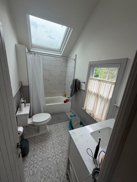 Bright modern bathroom with skylight, white subway tile shower, toilet, vanity sink, patterned floor, and window with natural light