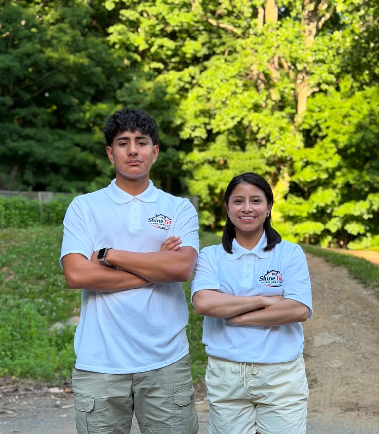 Two young people in matching light blue polo shirts standing together outdoors with arms crossed, smiling at the camera with green foliage in the background