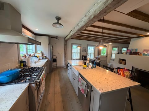 Spacious kitchen with gas stove, large island with cork countertop, exposed wooden beams, and multiple windows providing natural light