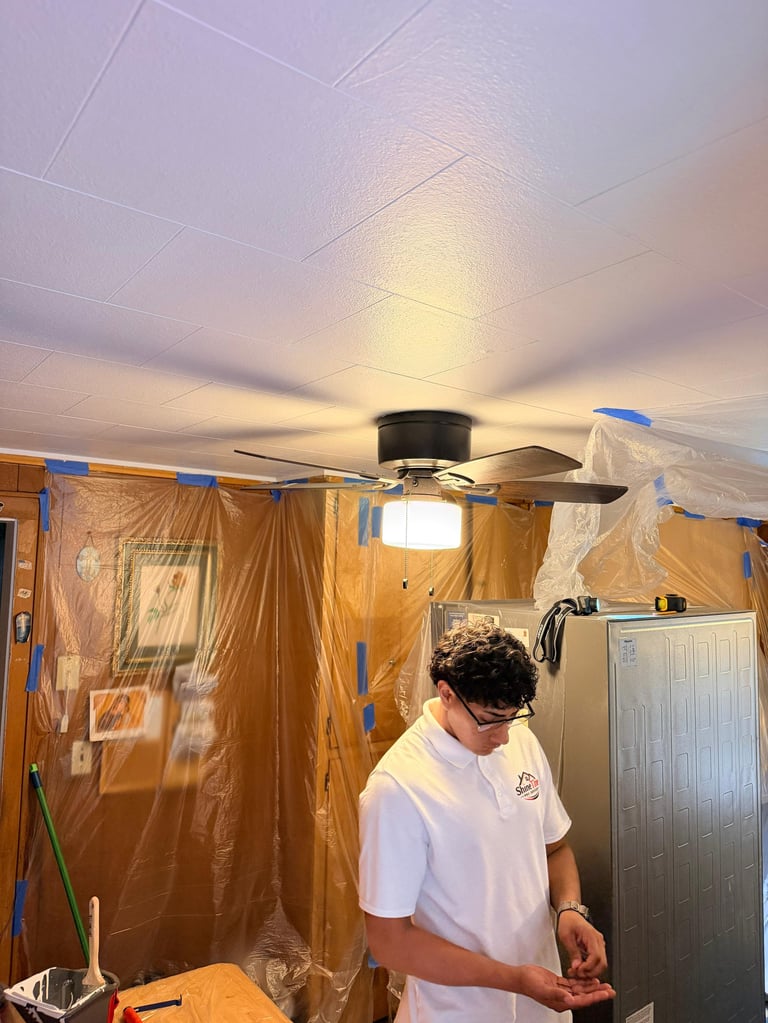 Worker in white shirt installing ceiling fan in room covered with protective plastic sheeting and blue tape