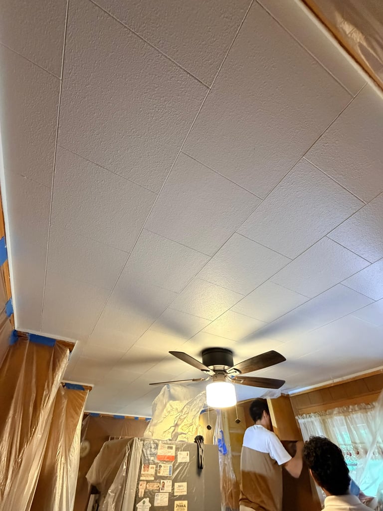 Modern ceiling fan with light fixture mounted on white drop ceiling in interior room with wooden trim and two people visible.
