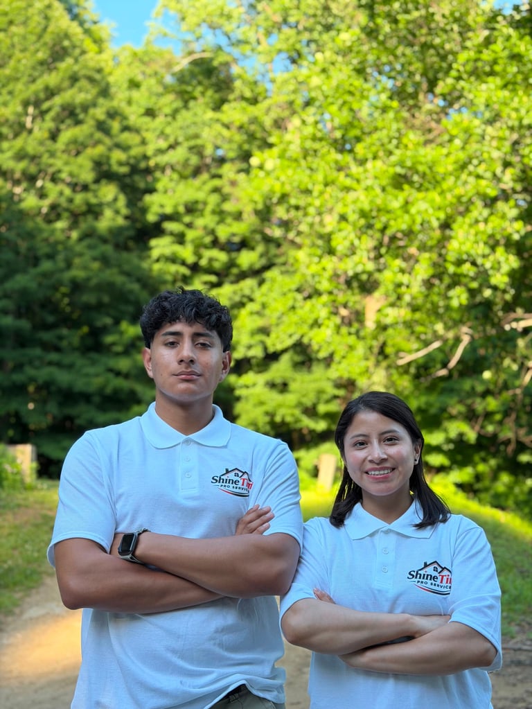 Two young people in matching light blue ShimFit polo shirts standing outdoors with arms crossed, green foliage and clear sky in background