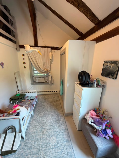 Bedroom interior with exposed wooden beams, white wardrobe, single bed with pink items, dresser, and window with air conditioning unit