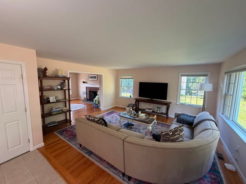 Spacious living room with curved beige sofa, flat-screen TV on stand, wooden shelving, hardwood flooring, and large windows with natural light
